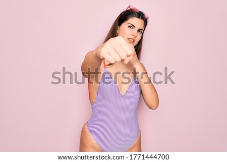 Young beautiful fashion girl wearing swimwear swimsuit and sunglasses over pink background Punching fist to fight, aggressive and angry attack, threat and violence