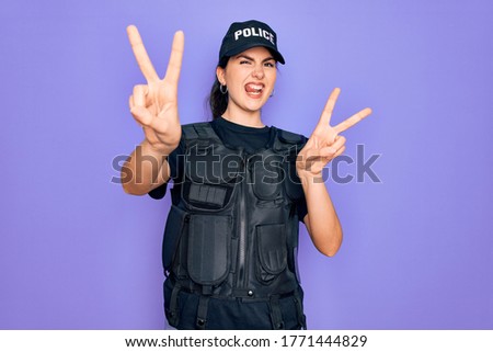 Young police woman wearing security bulletproof vest uniform over purple background smiling with tongue out showing fingers of both hands doing victory sign. Number two.