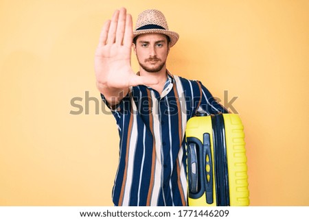 Young handsome man wearing summer hat holding cabin bag with open hand doing stop sign with serious and confident expression, defense gesture 