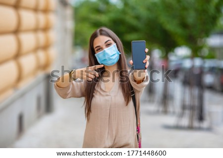 Woman wearing protective mask showing smart phone blank screen. Woman wearing protective face mask, showing mobile phone with blank white screen while standing in the city, Covid-19 concept. 