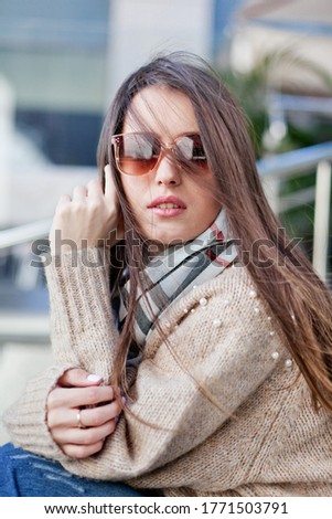 A brown-haired girl in casual style with a scarf around her neck and a light sweater sits on the steps. The average plan. Natural daylight.
