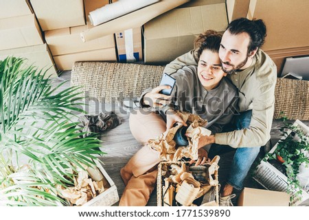 Lovely young husband and wife in casual clothes make selfie near pot palm tree and boxes with belongings waving hands in new house upper view.