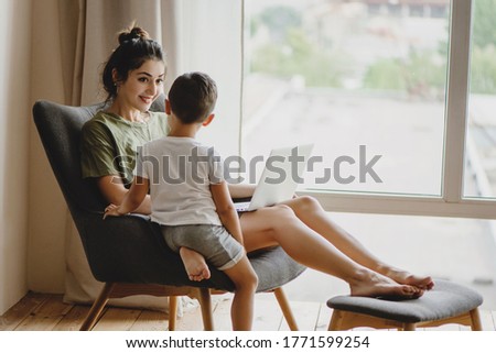 Young mother and her son sit together on chair with laptop.