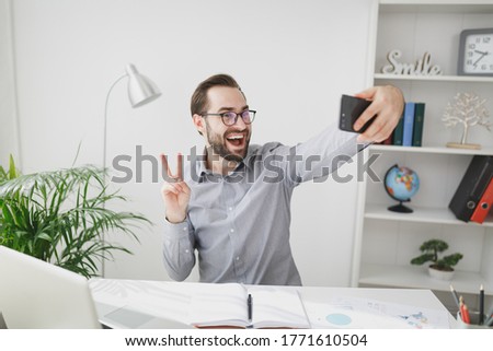 Cheerful business man in gray shirt sit at desk work on laptop in light office on white wall background. Achievement business career concept. Doing selfie shot on mobile phone, showing victory sign