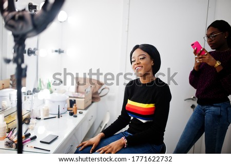 African American woman applying make-up by make-up artist at beauty saloon. Artist make photo on mobile phone of her work.