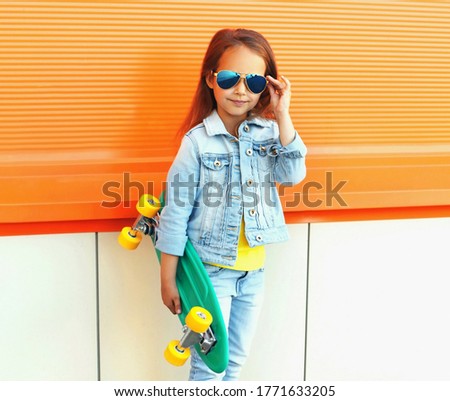 Stylish little girl child with skateboard on city street over orange wall background