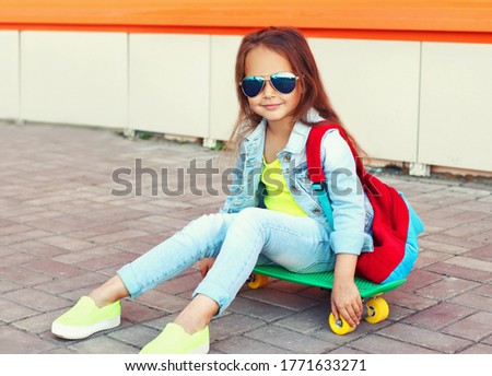 Portrait of little girl child sitting on skateboard with backpack on city street