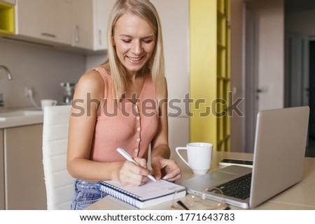 Smiling woman writing notes, studying online at home, sitting at home in the kitchen, happy girl wearing glasses watching webinar, listening course, learning language, making conference call