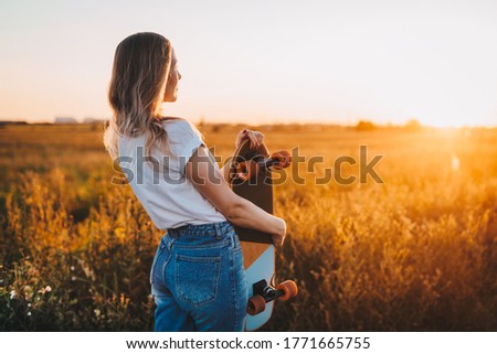a girl holds a longboard, a skateboard and looks at the sunset