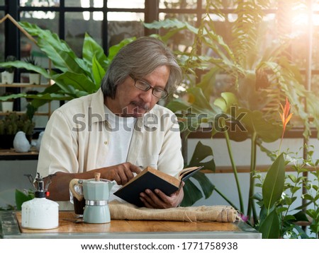 Healthy Asian senior man reading book while drinking hot coffee, sitting in the greenhouse. Happiness at home when staying at home. Elderly lifestyle.