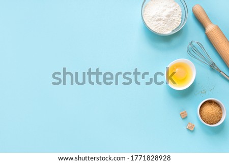 Baking utensils and ingredients.  Eggs, flour, sugar, whisk and rolling pin on blue background. Baking concept flat lay, copy space.