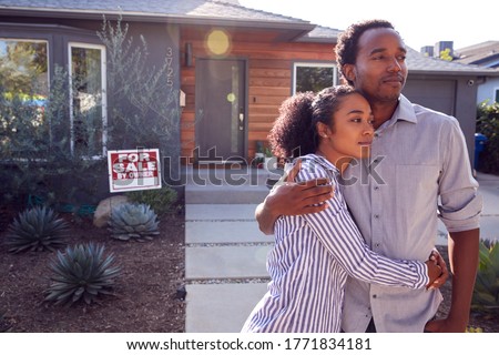 Thoughtful Couple Standing Outdoors In Front Of House With For Sale Sign In Garden