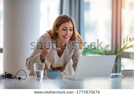 Woman sits at her desk checking her computer whilst eating a cornflakes from a bowl. She is wearing a smart trouser suit. Working through lunch. Female Worker In Office Having Healthy Lunch At Desk