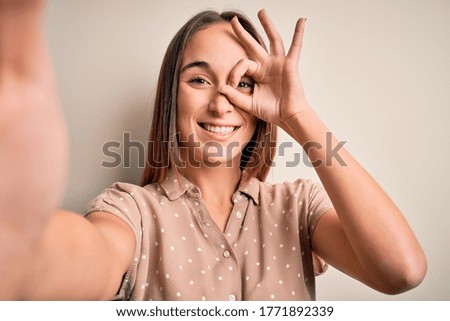 Young beautiful woman wearing summer shirt making selfie by camera over white background with happy face smiling doing ok sign with hand on eye looking through fingers