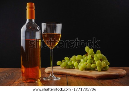 Still life with white juicy grapes, bottle of white wine without any label, and one wine glass. Dark black background, Wooden table.