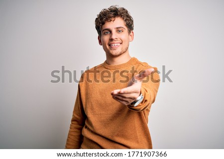 Young blond handsome man with curly hair wearing casual sweater over white background smiling cheerful offering palm hand giving assistance and acceptance.