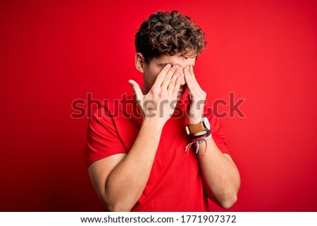 Young blond handsome man with curly hair wearing casual t-shirt over red background rubbing eyes for fatigue and headache, sleepy and tired expression. Vision problem