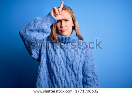 Young beautiful blonde woman wearing casual turtleneck sweater over blue background making fun of people with fingers on forehead doing loser gesture mocking and insulting.