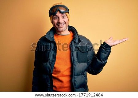 Young handsome skier man skiing wearing snow sportswear using ski goggles smiling cheerful presenting and pointing with palm of hand looking at the camera.