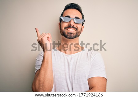 Young handsome man with beard wearing funny thug life sunglasses over white background with a big smile on face, pointing with hand and finger to the side looking at the camera.