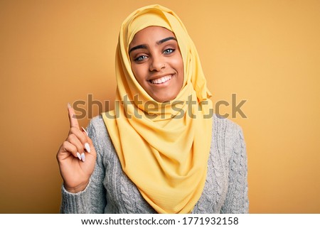 Young beautiful african american girl wearing muslim hijab over isolated yellow background with a big smile on face, pointing with hand finger to the side looking at the camera.