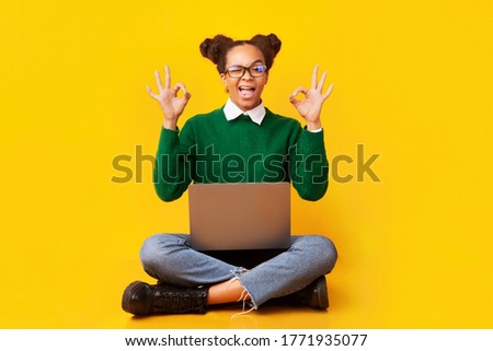I'm Fine. Excited afro teenage girl showing ok sign, sitting on floor with laptop over yellow background, copyspace