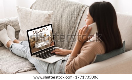 Asian girl booking hotel online, talking on phone with reservation team, sitting on couch at home at using laptop Royalty-Free Stock Photo #1771935086