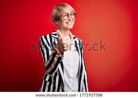 Blonde business woman with short hair wearing glasses and striped jacket over red background doing happy thumbs up gesture with hand. Approving expression looking at the camera showing success.