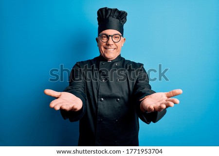 Middle age handsome grey-haired chef man wearing cooker uniform and hat smiling cheerful with open arms as friendly welcome, positive and confident greetings