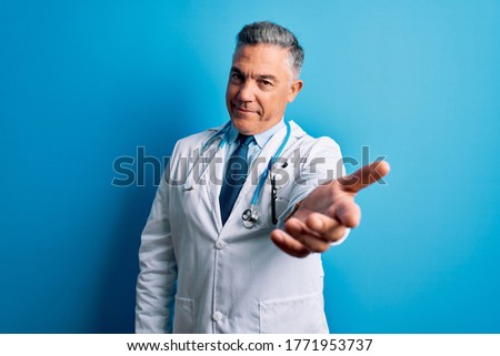 Middle age handsome grey-haired doctor man wearing coat and blue stethoscope smiling cheerful offering palm hand giving assistance and acceptance.