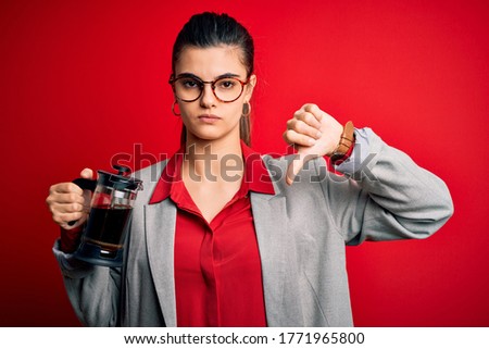 Young beautiful brunette woman doing coffe holding french coffeemaker over red background with angry face, negative sign showing dislike with thumbs down, rejection concept
