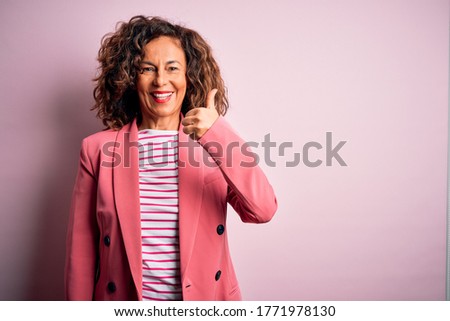 Middle age beautiful businesswoman wearing elegant jacket over isolated pink background doing happy thumbs up gesture with hand. Approving expression looking at the camera showing success.
