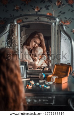 Beautiful long- and red- haired mother hugging her cute baby girl and sitting at the dressing table full of jewels. Image with selective focus and toning