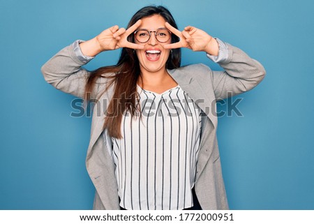 Young hispanic business woman wearing glasses standing over blue isolated background Doing peace symbol with fingers over face, smiling cheerful showing victory