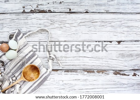 Apron with baking supplies. Whisk, measuring spoons, old wooden spoon and eggs over a white wood background. Image shot from top view.