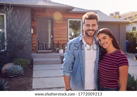 Portrait Of Couple Standing Outdoors In Front Of House With For Sale Sign In Garden Royalty-Free Stock Photo #1772009213