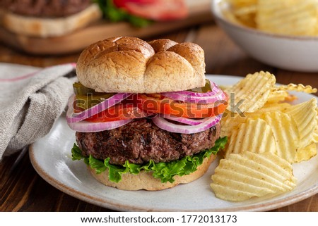 Juicy grilled hamberger with lettuce, tomato, pickle and onions on whole grain bun and potato chips on a plate