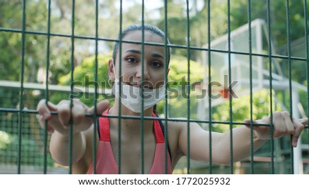 Female athlete behind wire fences. She smiles at the camera. She wears a medical mask to protect it from the virus.