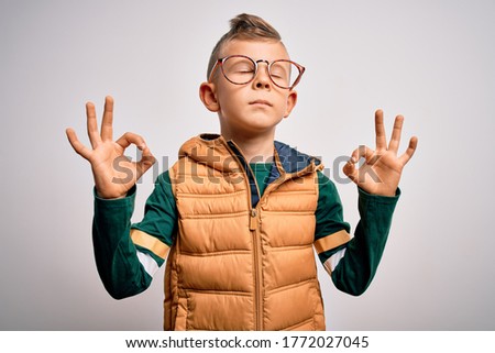 Young little caucasian kid with blue eyes wearing winter coat and smart glasses relax and smiling with eyes closed doing meditation gesture with fingers. Yoga concept.