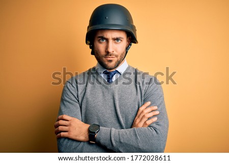Young handsome businessman wearing military helmet over isolated yellow background skeptic, disapproving expression on face with crossed arms. Negative person.