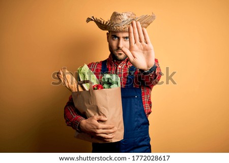 Young rural farmer man holding fresh groceries from marketplace over yellow background with open hand doing stop sign with serious and confident expression, defense gesture