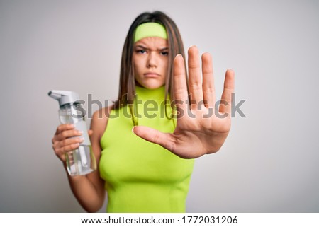 Beautiful sporty girl doing sport wearing sportswear drinking bottle of water to refressment with open hand doing stop sign with serious and confident expression, defense gesture