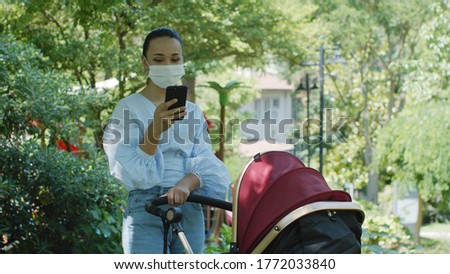 A young mother in a summer dress is looking at the phone with her other hand holding a stroller. A walk with a stroller in the park.