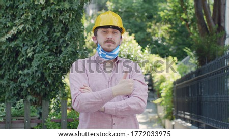 Portrait of male engineer with medical mask with Greece flag and yellow helmet. Serious. Everything is OK sign.