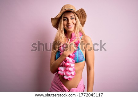 Young beautiful blonde woman on vacation wearing bikini and hat with hawaiian lei flowers cheerful with a smile of face pointing with hand and finger up to the side with happy and natural expression