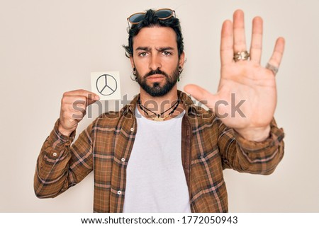 Young handsome hispanic bohemian hipster man with bear holding paper note with peace symbol with open hand doing stop sign with serious and confident expression, defense gesture