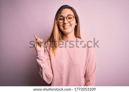Beautiful blonde woman with blue eyes wearing sweater and glasses over pink background with a big smile on face, pointing with hand and finger to the side looking at the camera.
