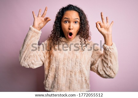 Young beautiful woman with curly hair wearing casual sweater standing over pink background looking surprised and shocked doing ok approval symbol with fingers. Crazy expression