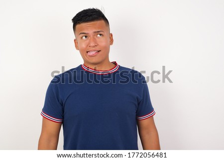 Young handsome hispanic man wearing casual t-shirt standing over white isolated background showing grimace face crossing her eyes and showing tongue . Being funny and crazy