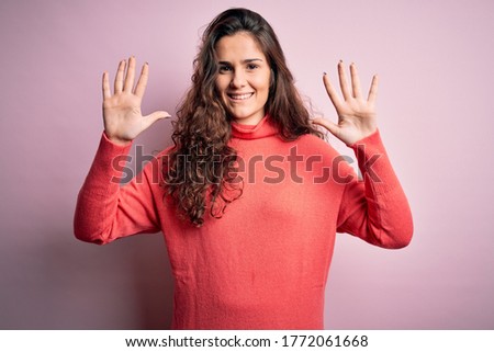 Young beautiful woman with curly hair wearing turtleneck sweater over pink background showing and pointing up with fingers number ten while smiling confident and happy.